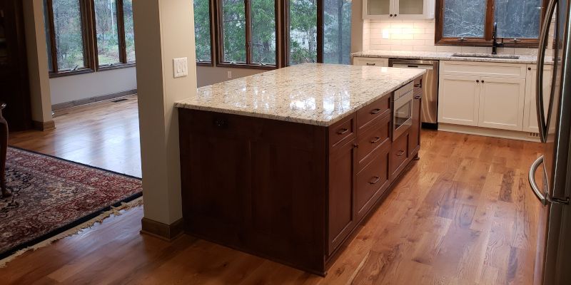 Modern kitchen with a large granite island, wood cabinets, hardwood floors, and windows overlooking greenery