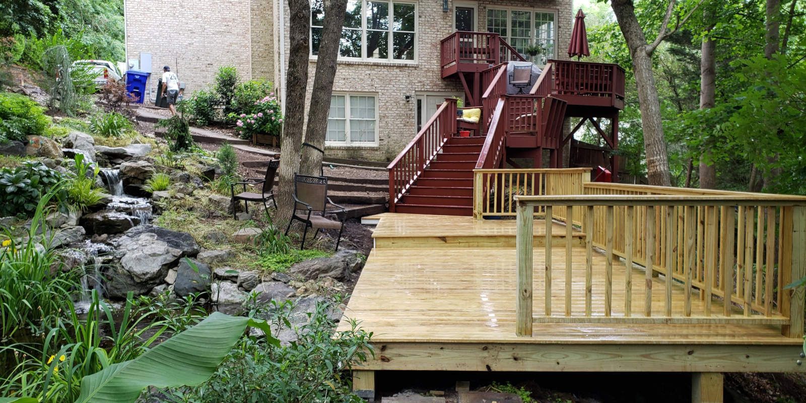 Backyard deck with red stairs beside a stone wall and stream, surrounded by lush greenery