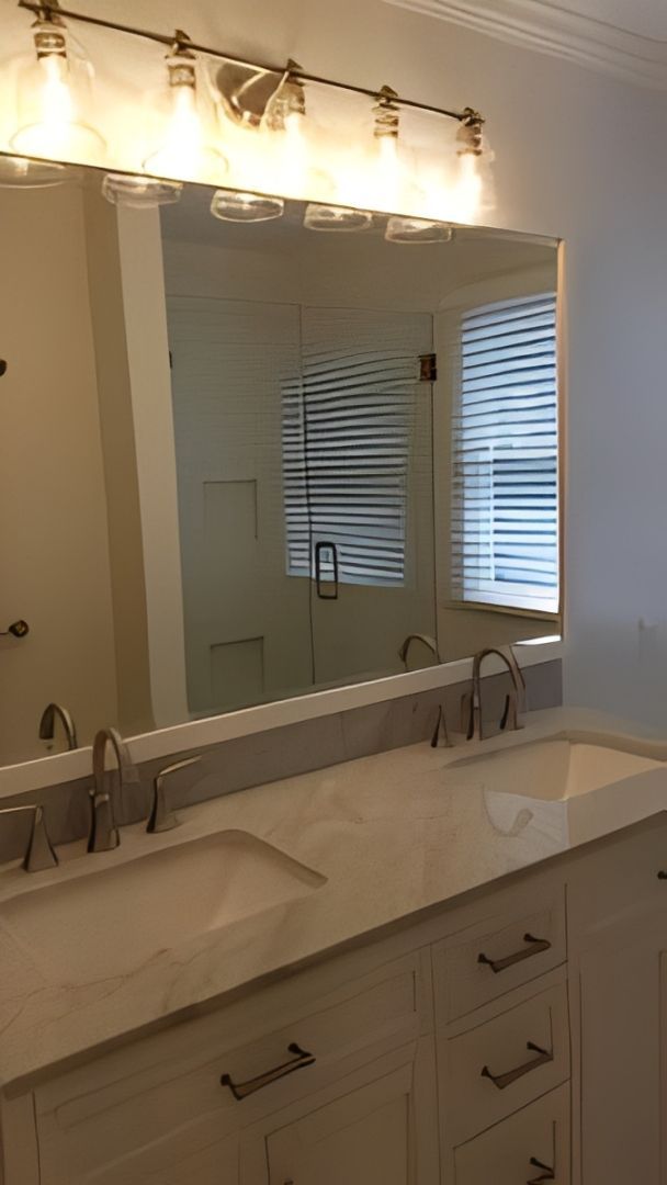 Bathroom vanity with double sinks, large mirror, and lit wall sconce above a white countertop.