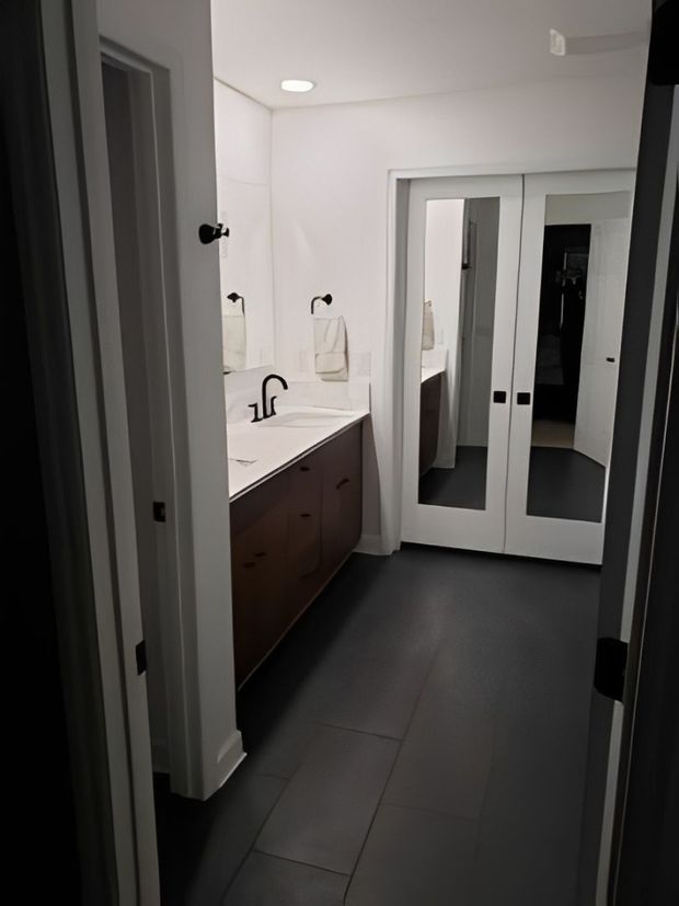 Narrow modern bathroom with white walls, dark floor tiles, vanity, sink, and mirrored double doors.