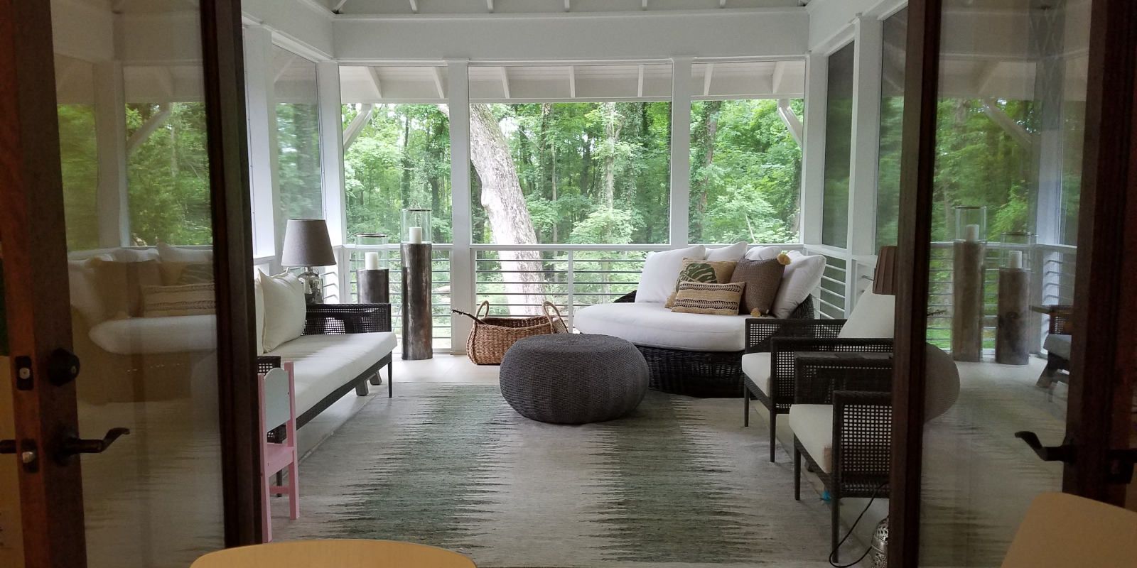 Bright sunroom with white sofas, patterned rug, wicker chairs, and large windows overlooking greenery