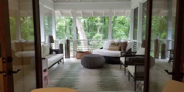 Bright sunroom with white sofas, rattan chairs, and lush green outdoor view through large windows
