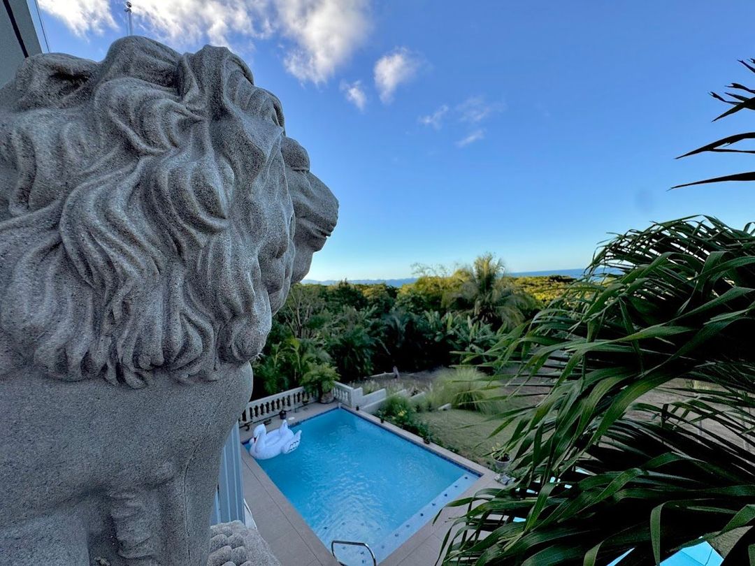 Lion statue overlooking a blue pool and tropical landscape under a partly cloudy sky.