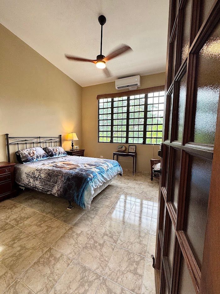 Bedroom with a bed, window, and ceiling fan. Light-colored walls and floor. View through an open door.