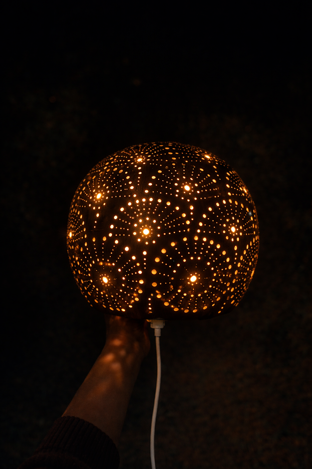 A dark, spherical, perforated lamp glowing with warm internal light, held by a person's hand against a black background.