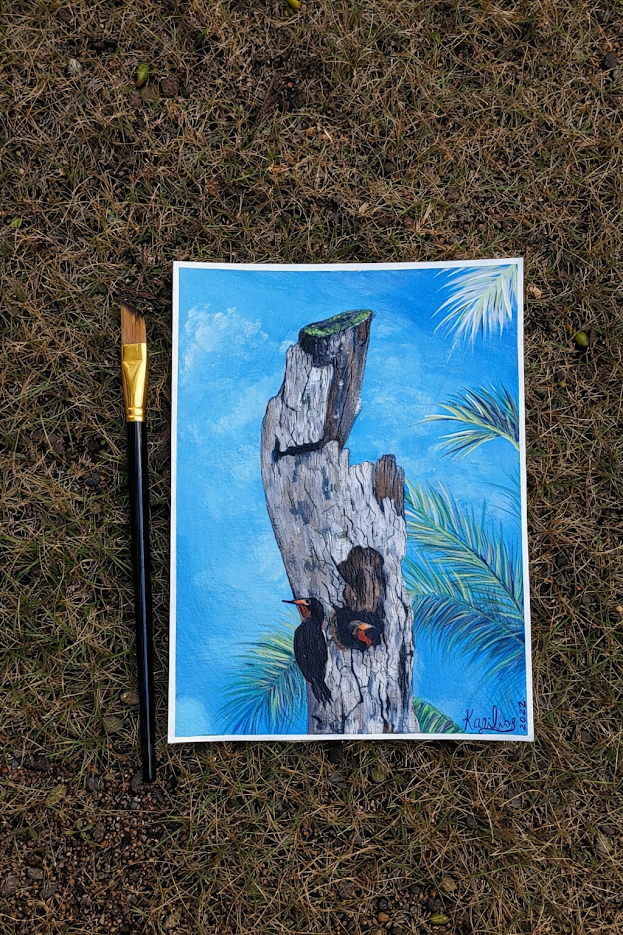 A painting of a woodpecker on a tree trunk against a blue sky, placed on grass next to a paintbrush.