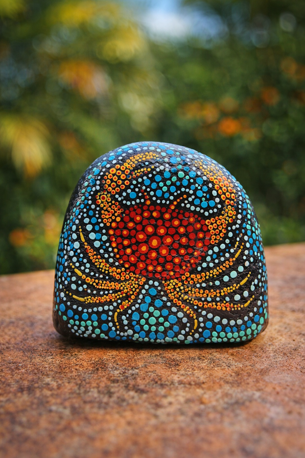A rock painted with a vibrant dot-art crab featuring a red body and teal legs, resting on a textured surface.
