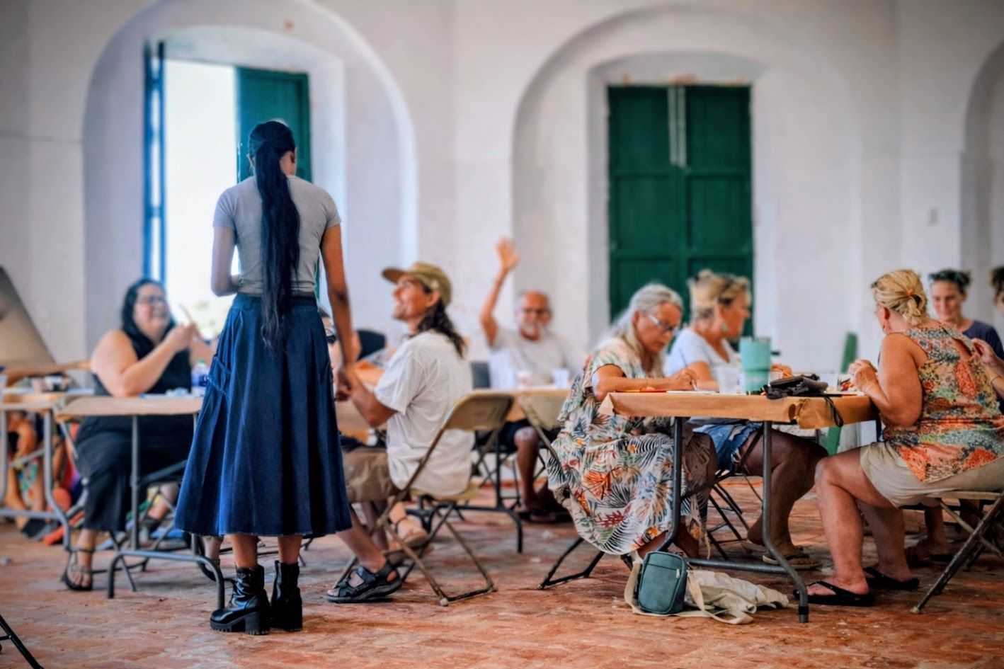 A group sits at tables in a bright, arched room as one person stands in the foreground, facing the group.