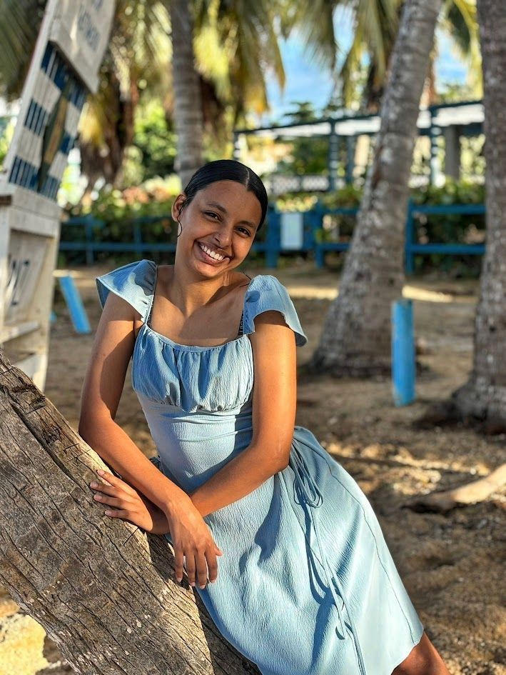 A person in a light blue sundress leaning against a tree trunk on a sandy beach with palm trees in the background.