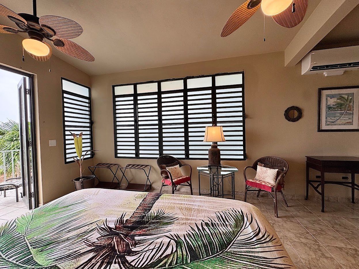 Bedroom with large windows, tropical bedding, wicker chairs, and ceiling fans.
