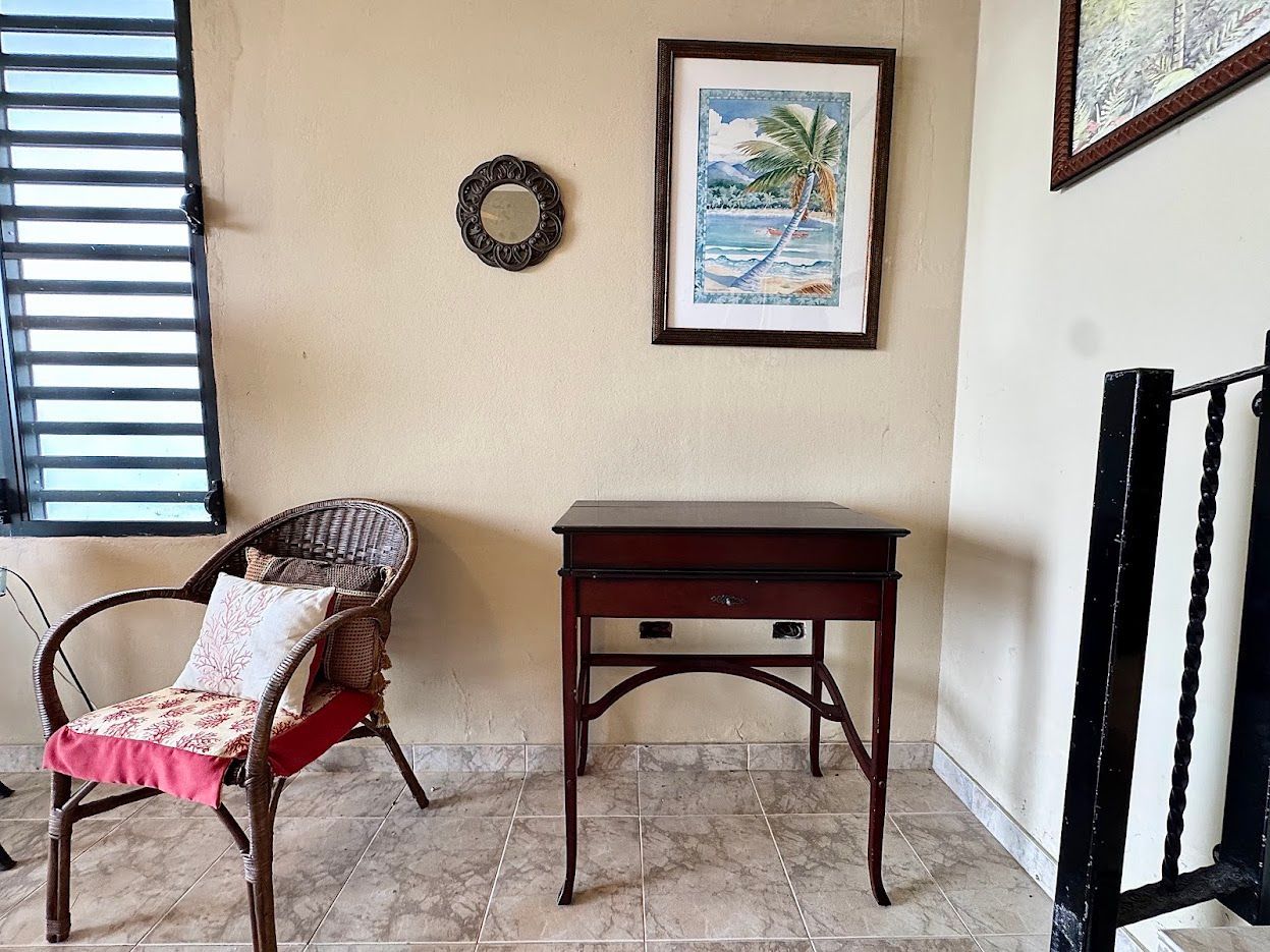 Wicker chair with pillow, small desk, and art on a beige wall next to a window with blinds.