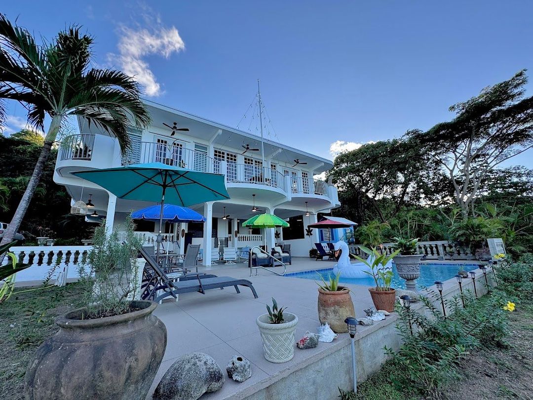 White house with pool, patio, and umbrellas under a blue sky, surrounded by tropical plants.