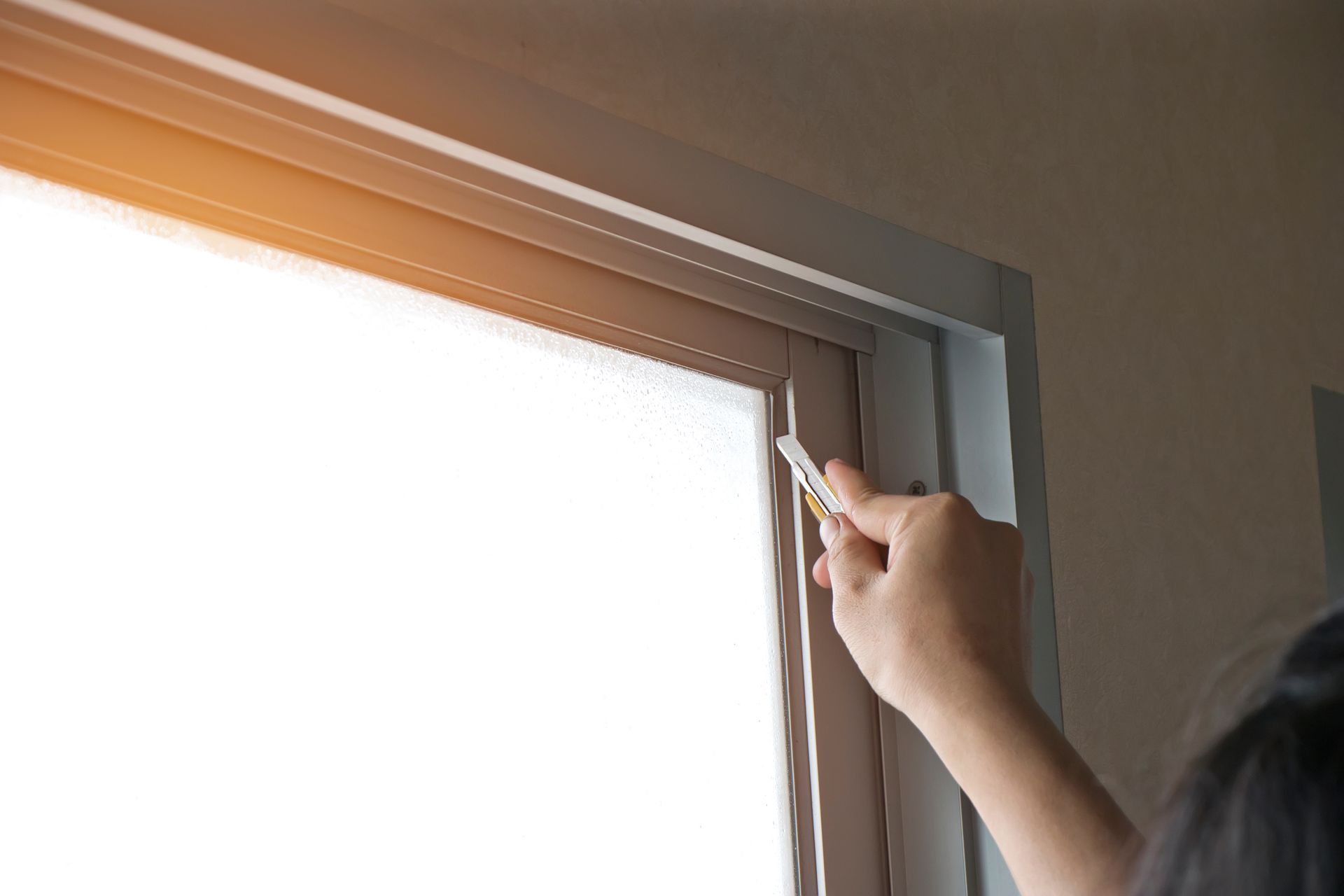 Hand using a utility knife to remove old caulk from a window frame.