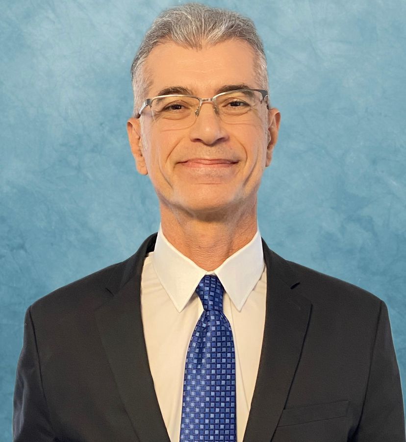 Man in a suit, wearing glasses, smiling, against a blue background.