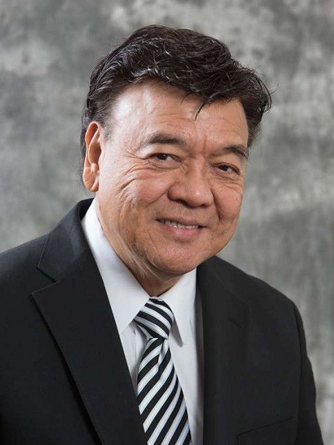 Man in a suit smiles, dark hair, striped tie, against a gray background.
