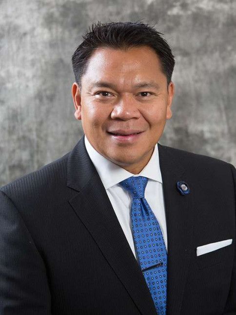 Man in a suit, smiling, with blue tie and white pocket square; against a gray backdrop.