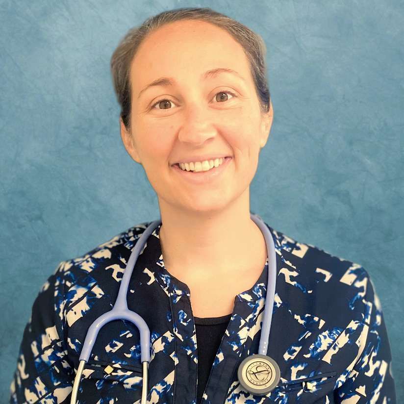 Woman with short hair, stethoscope, blue patterned jacket, smiling in front of a blue background.