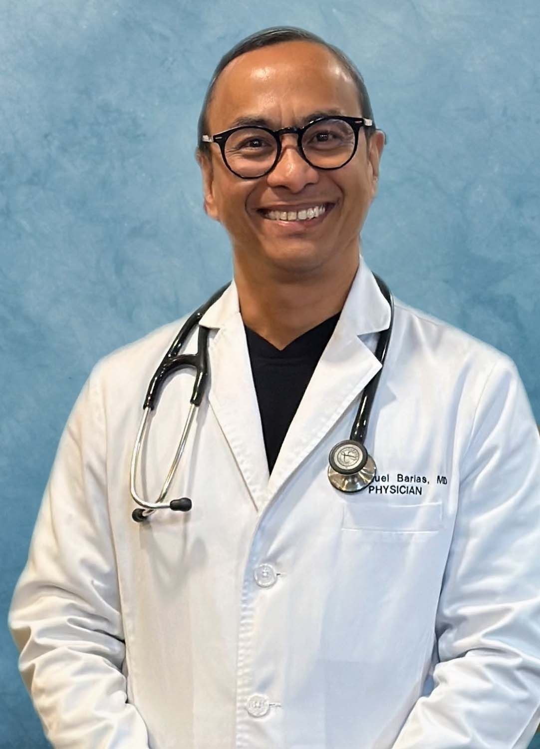 Doctor smiling, wearing glasses and stethoscope, in front of a blue background.