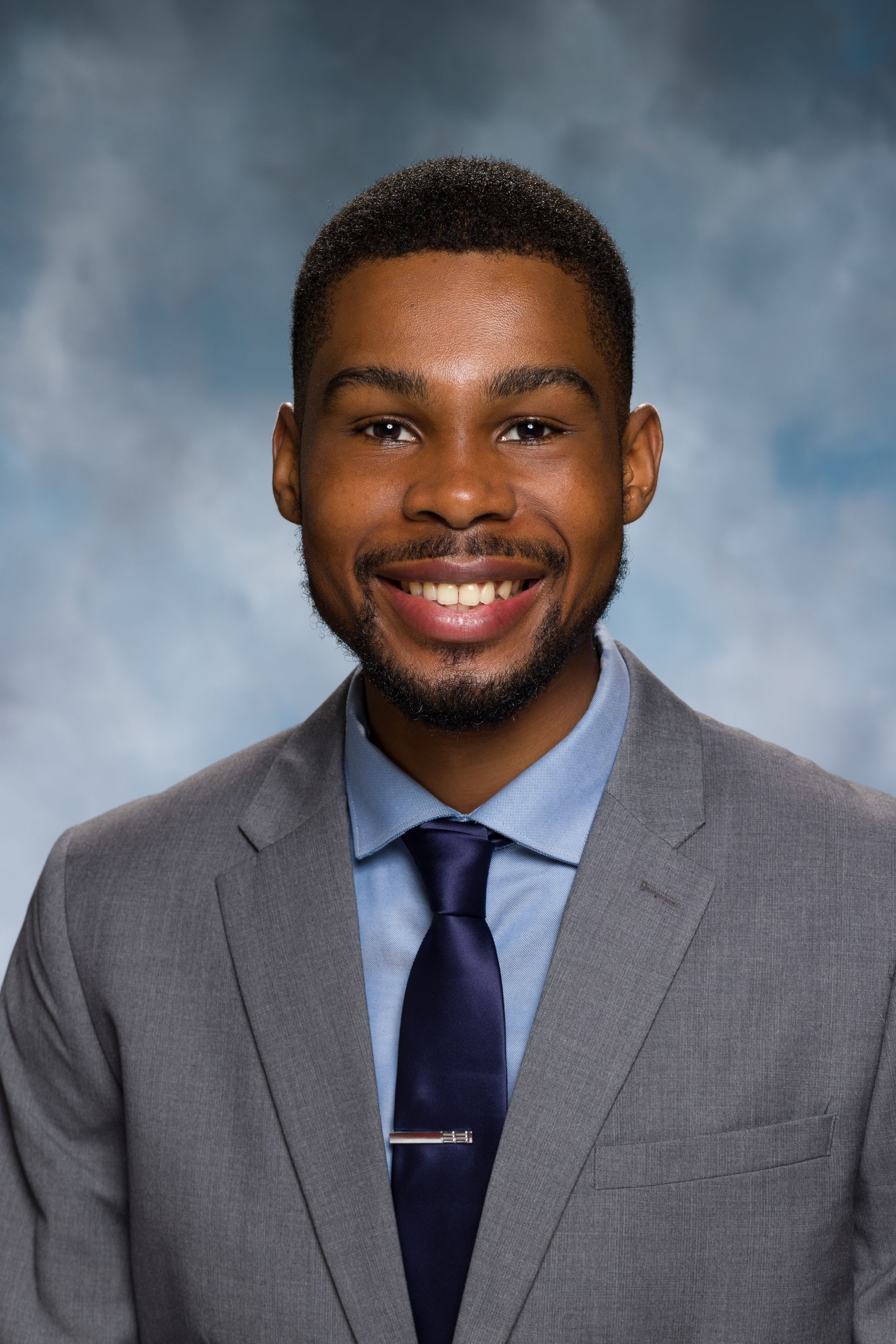 Man in gray suit smiles, dark blue tie, light blue shirt, against a cloudy backdrop.