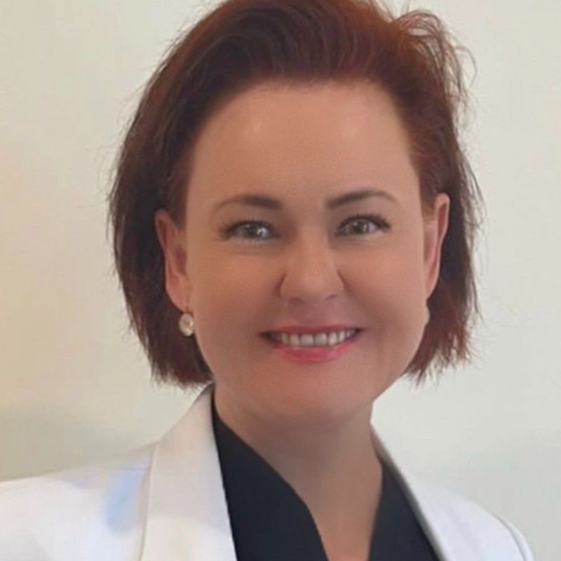 Woman with short auburn hair and a friendly smile, wearing a white blazer, against a light background.