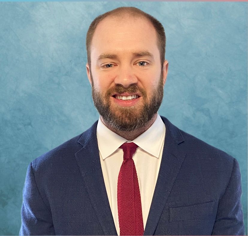 Man in a blue suit with red tie smiling in front of a blue background.