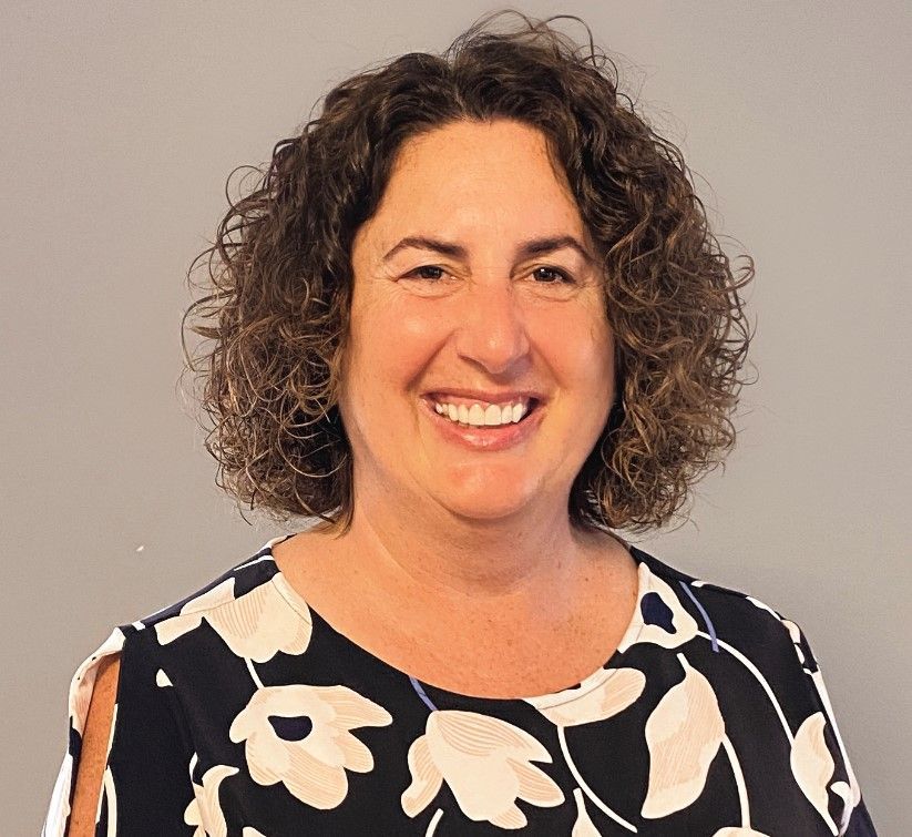 Woman with curly brown hair smiles broadly, wearing a black and white floral print top, against a gray background.