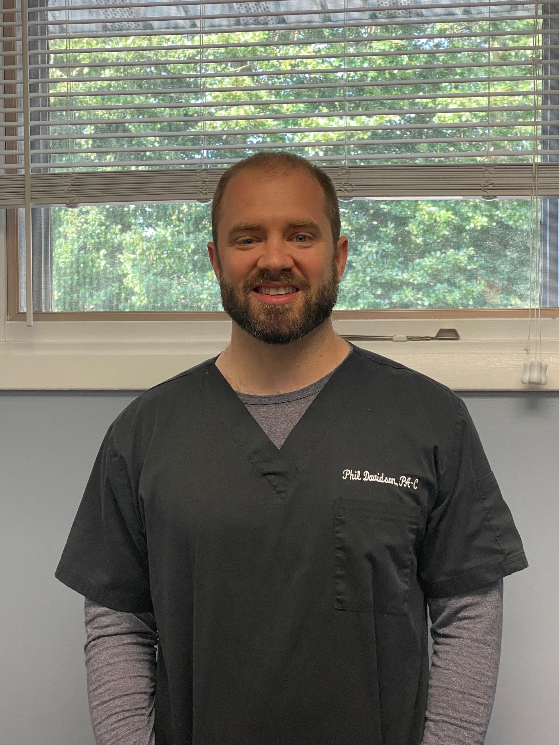 Man in scrubs smiles, standing indoors by a window with blinds.