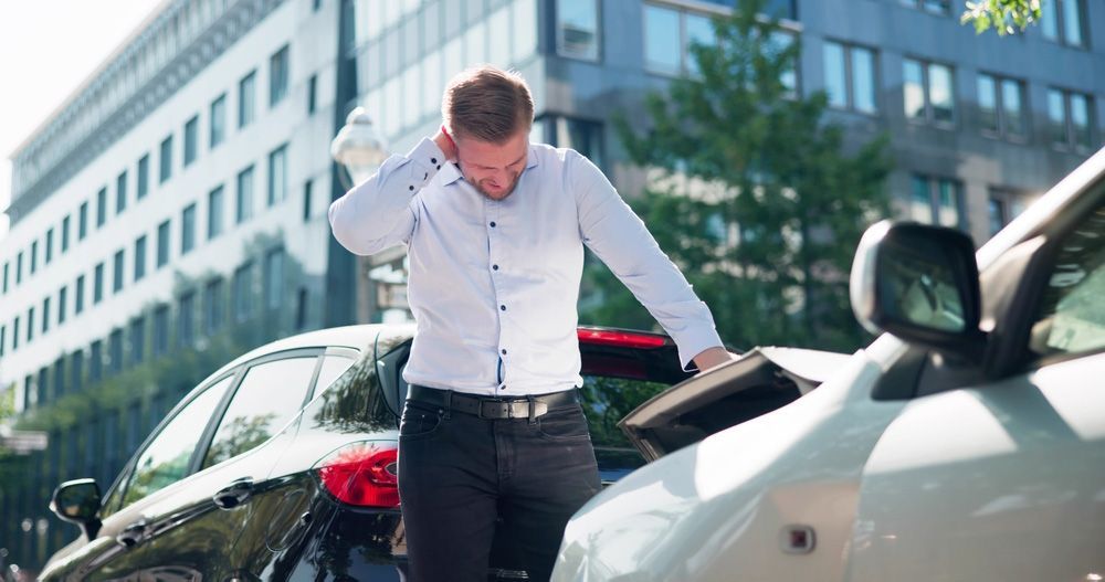 Man in white shirt standing between two cars on a city street, looking down at damage on one car