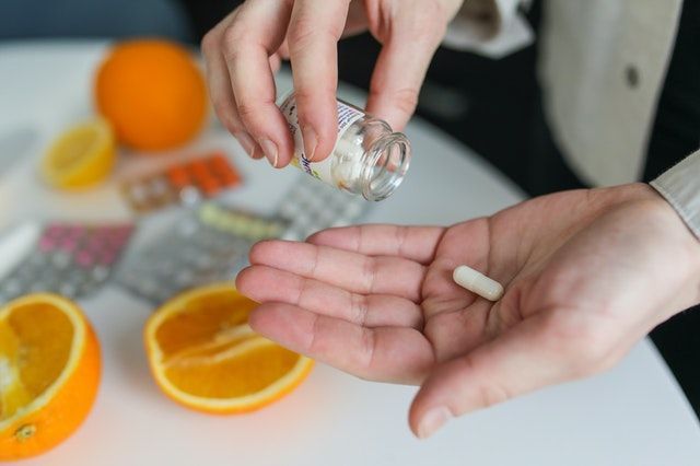 Person pouring pills from a bottle into their hand, near sliced oranges and medicine packets.