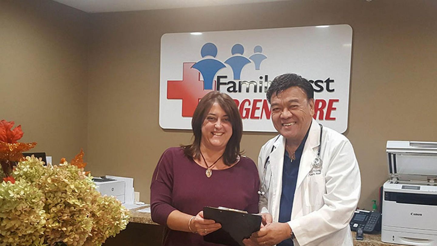 Woman and doctor at Family First Emergency Care, smiling and holding a clipboard. Sign with logo visible.