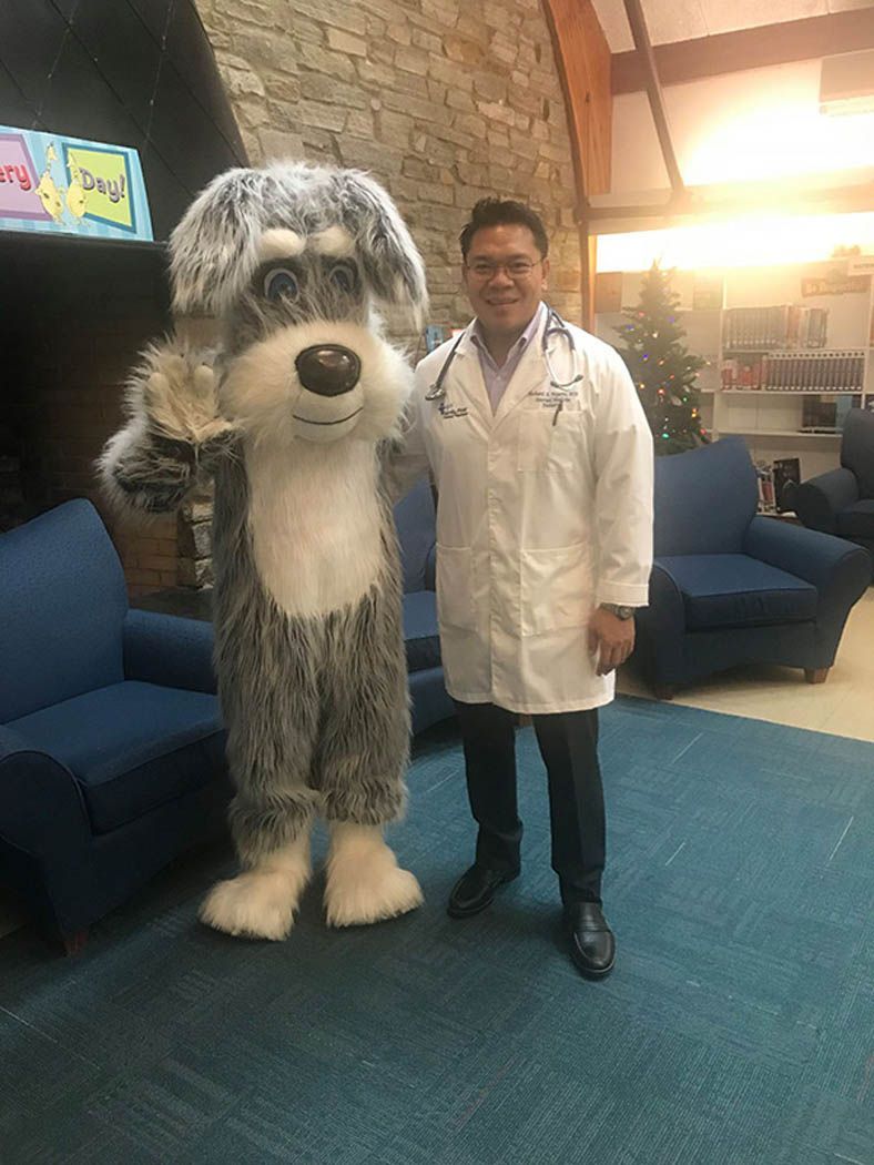 Doctor with stethoscope, smiles, poses with dog mascot in waiting room.