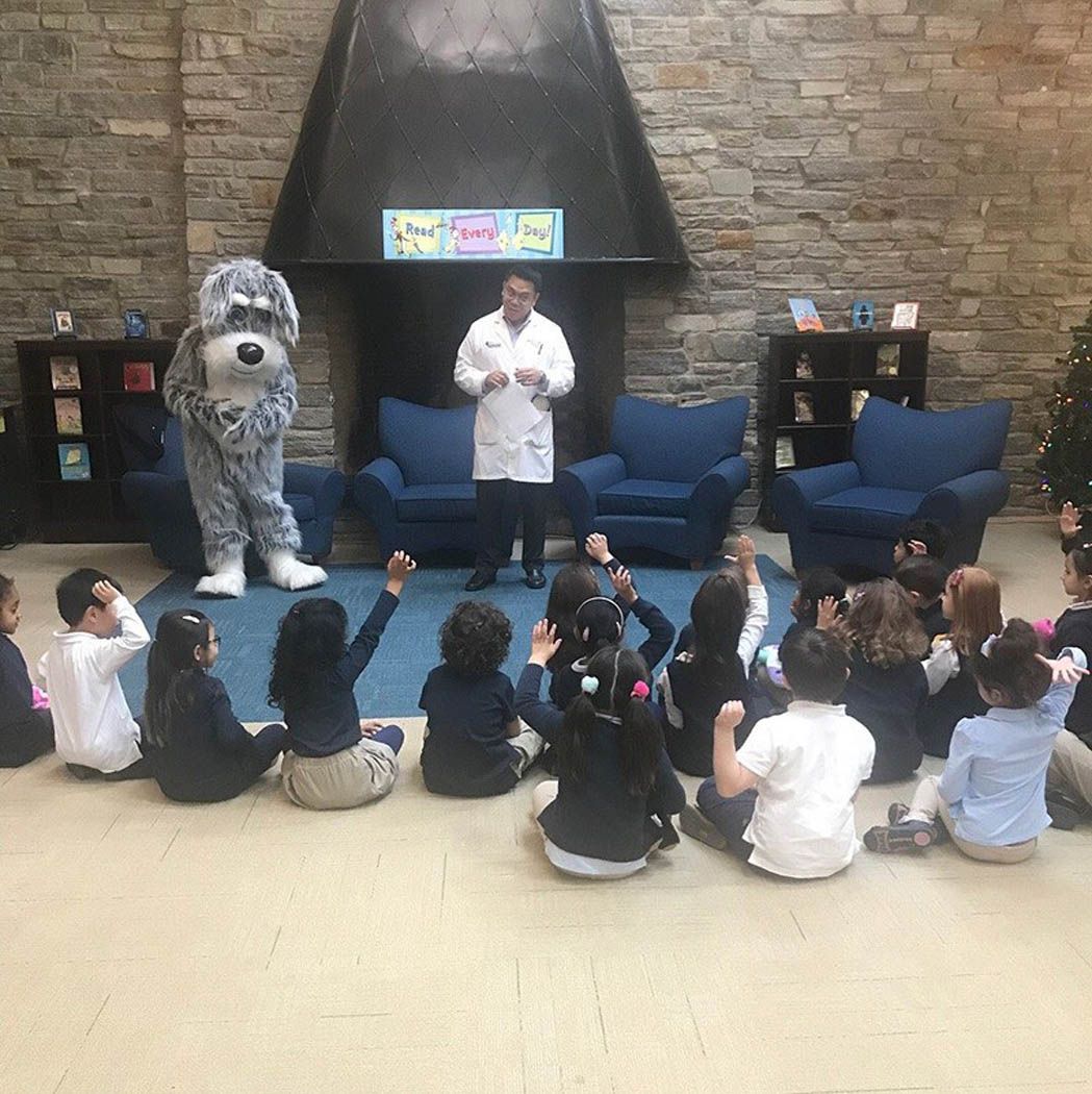 Children seated on floor listening to a man and a dog mascot in front of a fireplace.