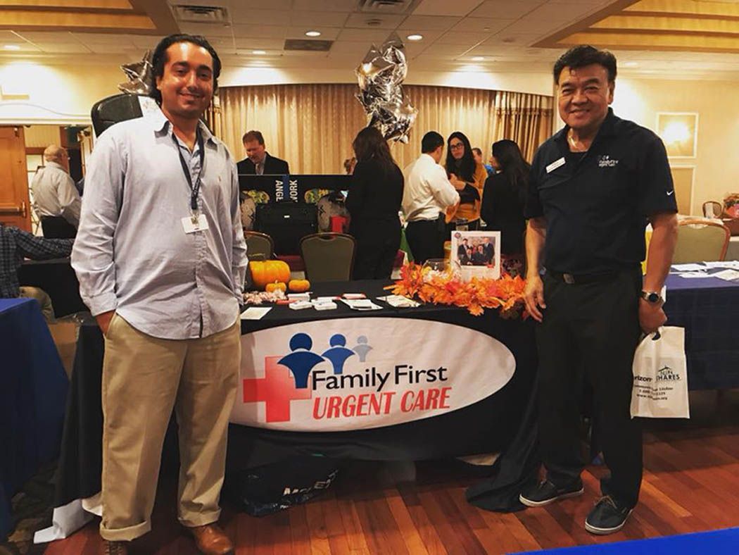 Two men stand at a Family First Urgent Care booth. They are at an event with other booths and people.