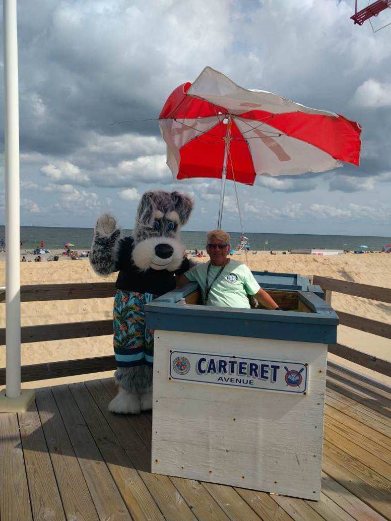 A mascot and person in a booth on a boardwalk in front of the beach. Mascot waves.