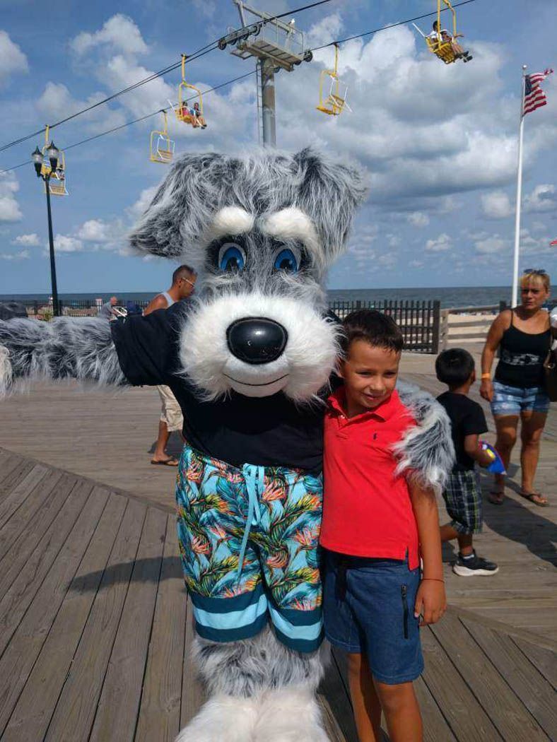 Boy poses with a dog mascot on a boardwalk. Both wear summery outfits. Sky and ocean in background.