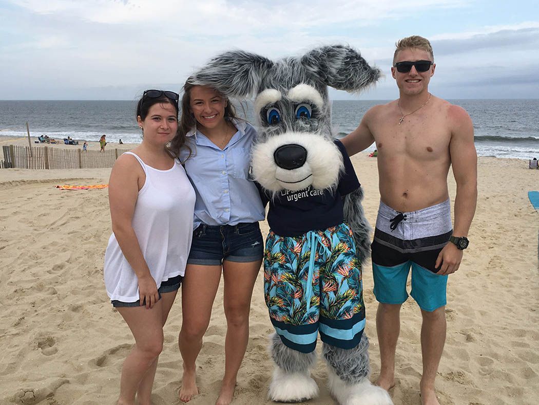 Four people on a beach: two women, a costumed dog, and a shirtless man, smiling at the camera.