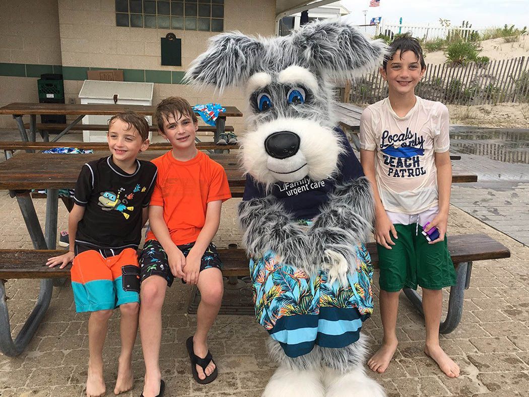 Three boys and a dog mascot pose on a bench outdoors. The boys wear swimwear, and the mascot has a blue scarf.