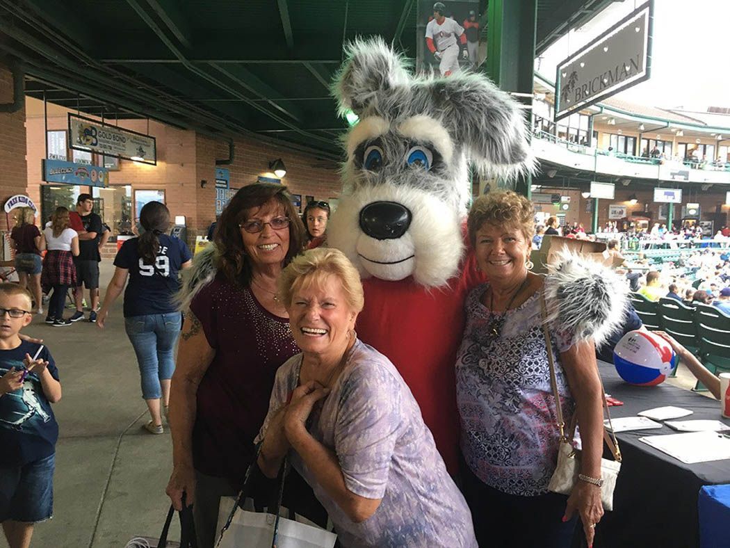 Three women pose with a dog mascot at a baseball stadium. People and seats are visible in the background.