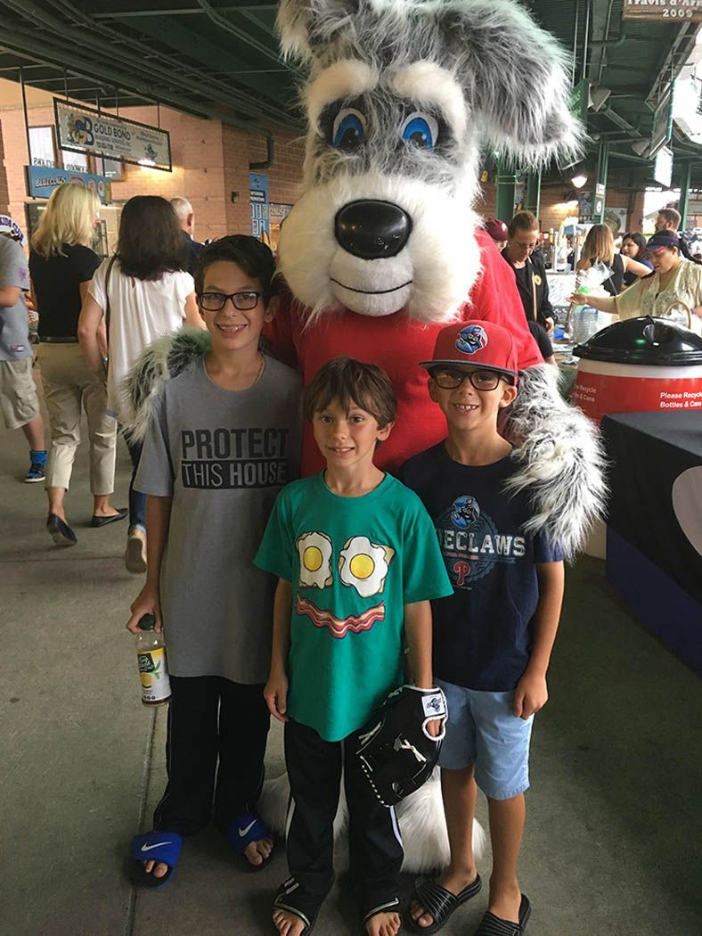 Three boys pose with a dog mascot at a baseball game.