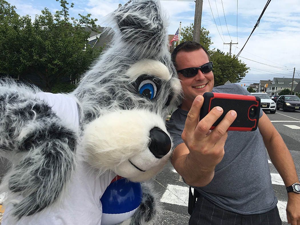 Man in sunglasses taking a selfie with a gray and white dog mascot on a street.