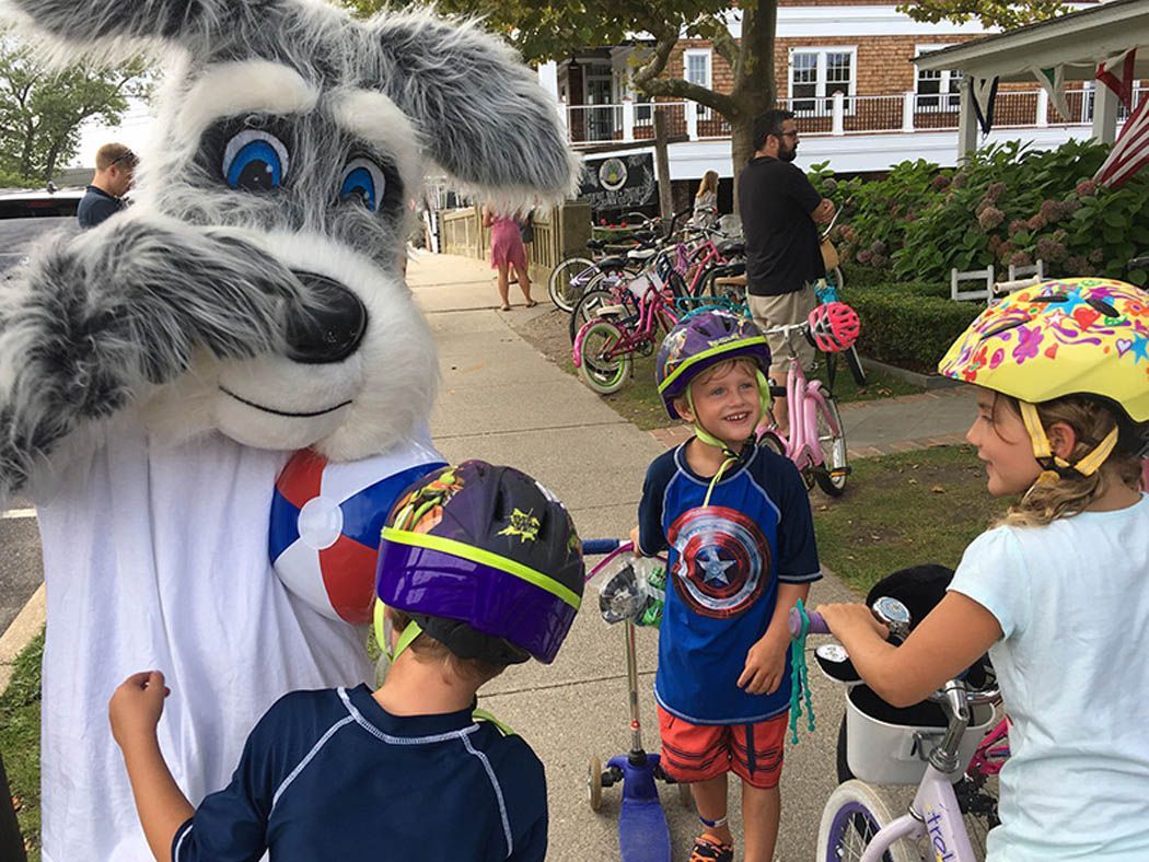 A mascot poses with two children in helmets, bikes in background on a sidewalk.