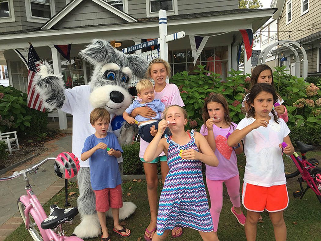 A mascot with children blowing bubbles in front of a house decorated with flags.