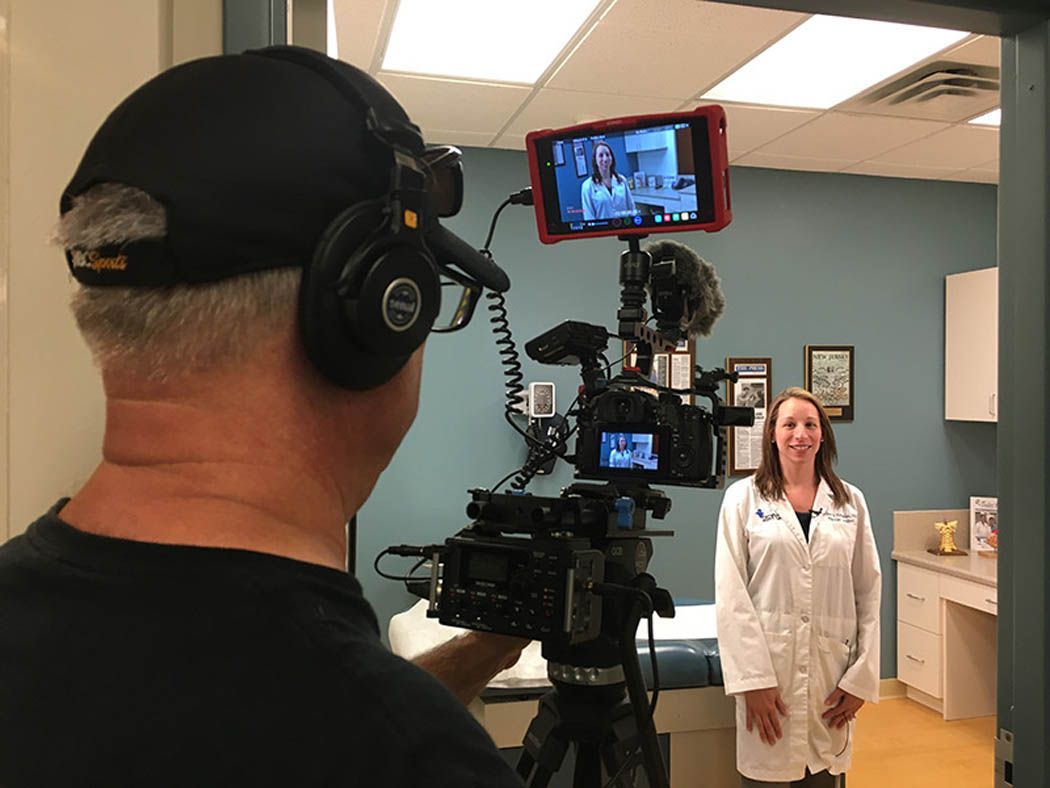 Videographer filming a woman in a white coat inside a medical office; blue wall background.