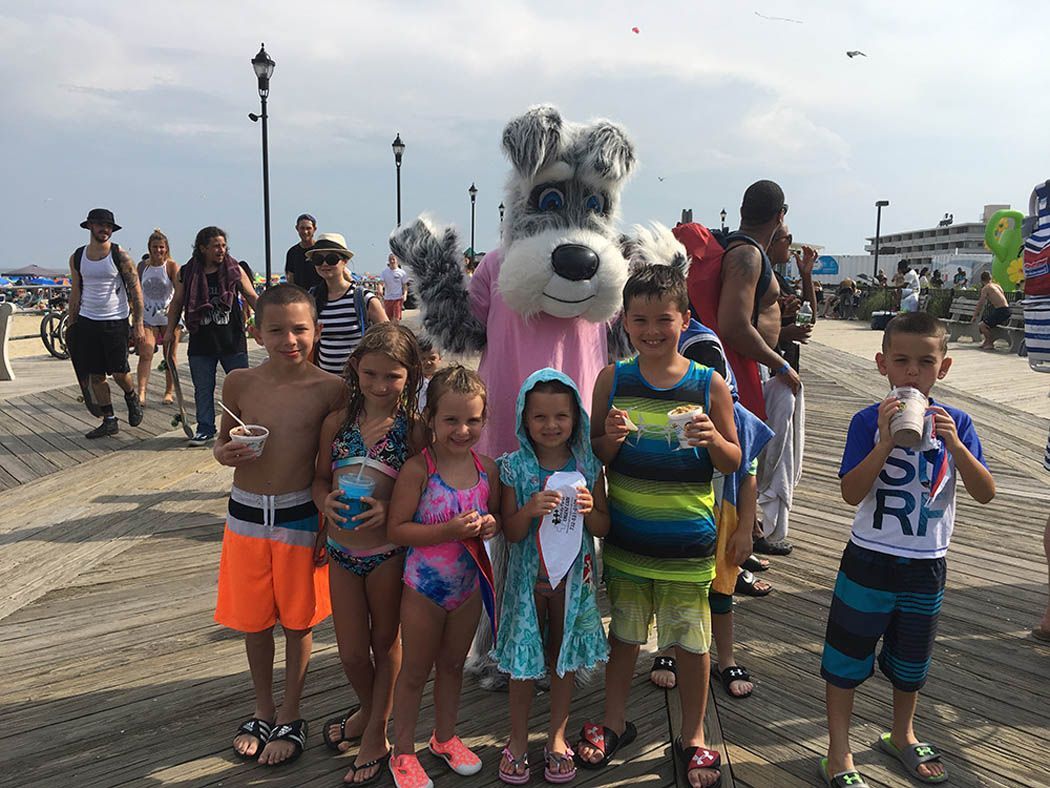 Children pose with a dog mascot on a boardwalk; some hold beverages.