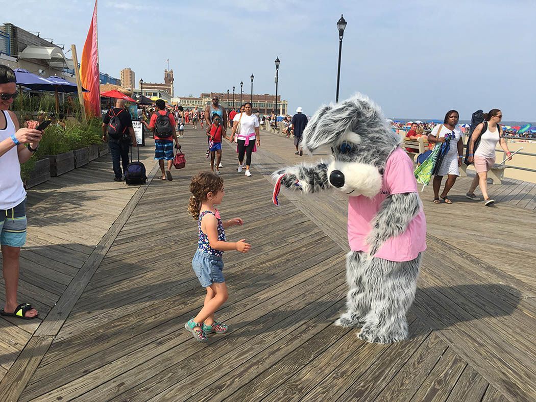 A girl running towards a costumed dog character on a boardwalk; people walking in the background.