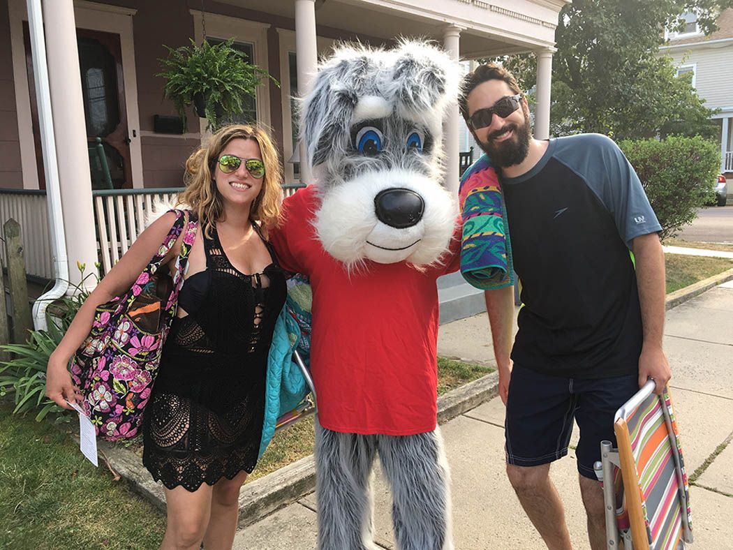 A woman and a man pose with a dog mascot on a sidewalk, smiling. The setting appears to be a residential area.