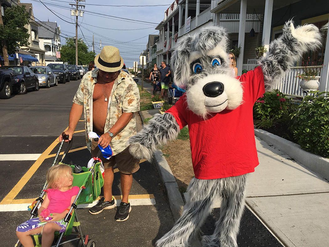A person in a dog mascot costume waves excitedly. A man pushes a stroller with a child on a street lined with parked cars.