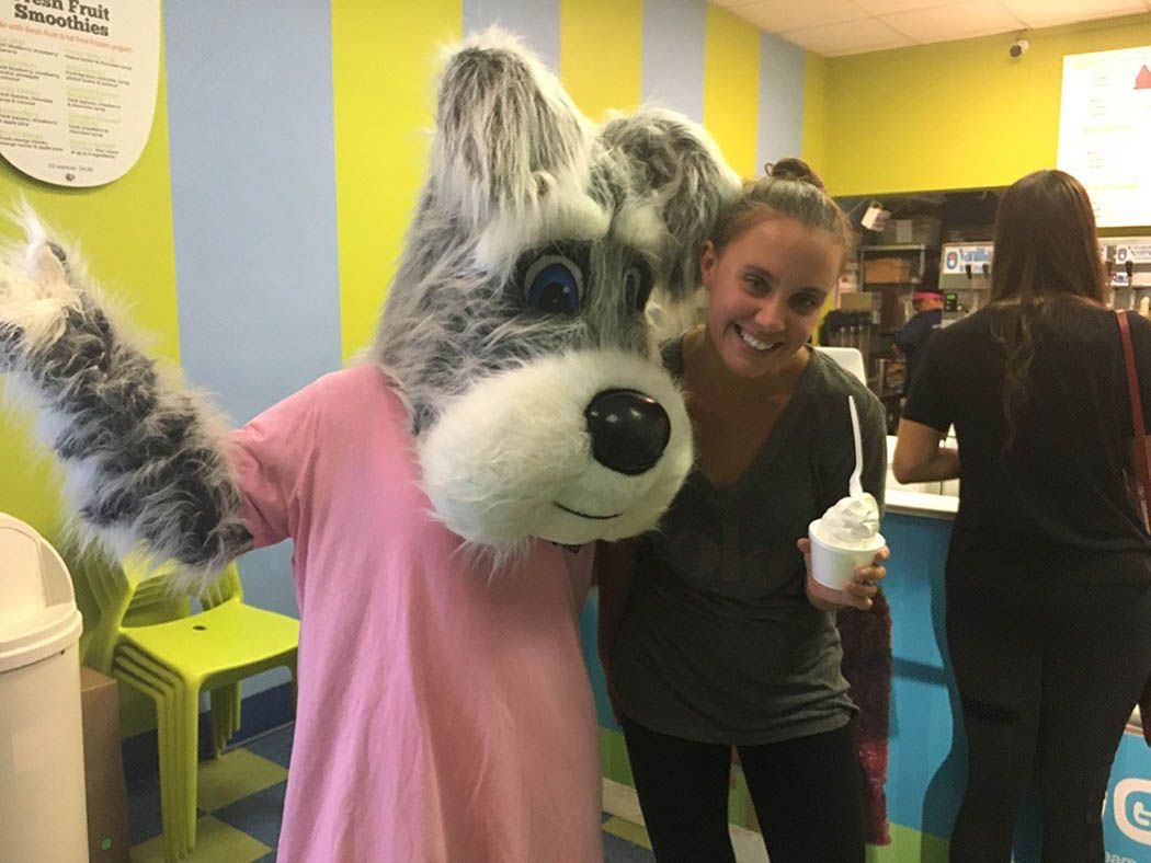 Woman smiles next to a gray and pink dog mascot, holding ice cream, in a brightly colored shop.