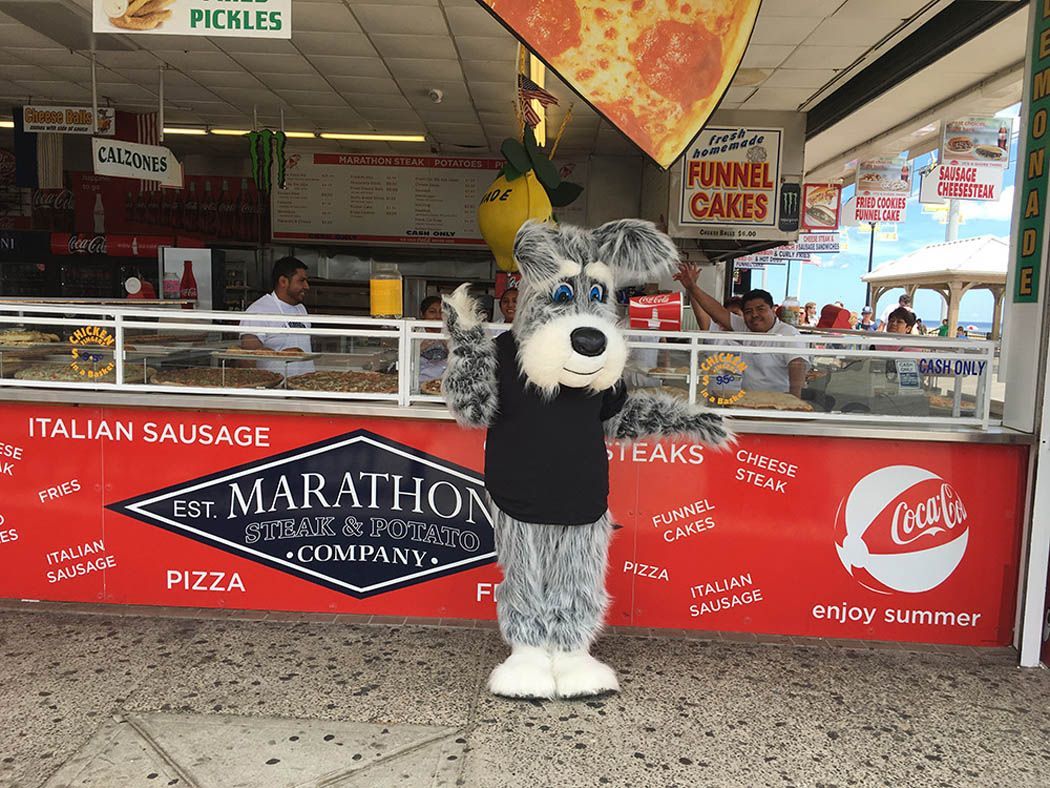 Dog mascot stands in front of a Marathon Pizza food stand. Arms outstretched. Red and white signage.