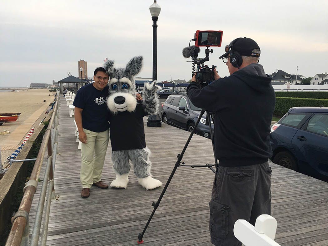 Man and mascot pose for video on a boardwalk, with a cameraman recording.